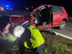 Czerwony rozbity samochód, policjant wykonujący oględziny pojazdu.