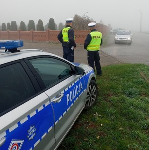 Radiowóz, dwóch policjantów ruchu drogowego stojący przed radiowozem, w tle mgła i pojazd.