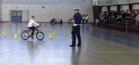 policjant z ruchu drogowego prowadzi egzamin na kartę rowerowa dla uczniów SP w poddębicach. Egzamin odbywa sie na sali gimnastycznej przy szkole. Obok policjanta dziecko manewruje rowerem omijając słupki.