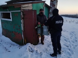 Na zdjęciu umundurowany funkcjonariusz policji, który przekazuje torby z odzieżą i żywnością osobie bezdomnej. W tle pustostan, śnieg i krzewy.