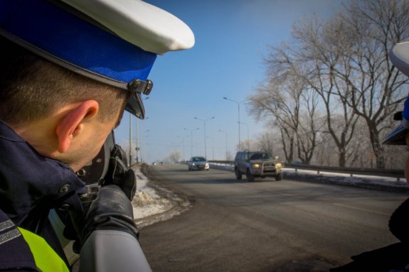 policjant w białej czapce stoi przy drodze i radarem mierzy prędkość pojazdów