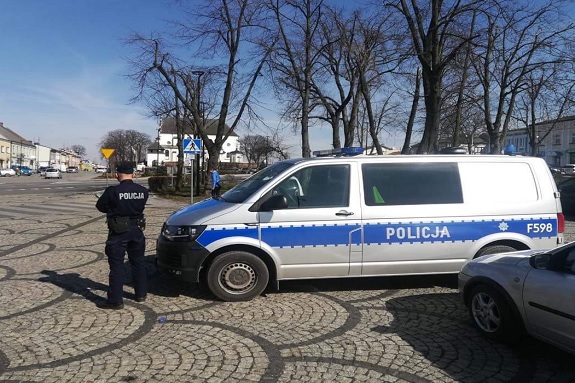 Na zdjęciu umundurowany funkcjonariusz policji, który stoi obok oznakowanego radiowozu. W tle inne pojazdy, znaki drogowe, drzewa, krzewy, budynki.