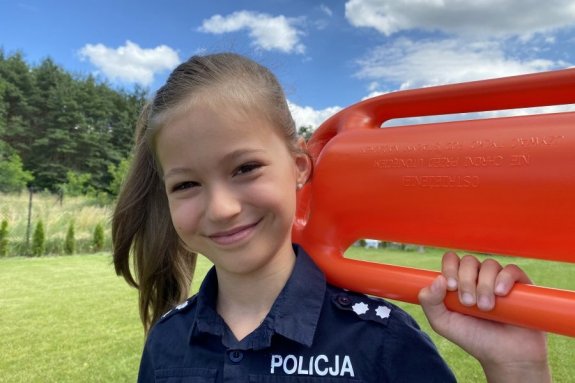 Na zdjęciu uśmiechnięta dziewczynka w koszuli granatowej z napisem policja, trzymająca bojkę ratowniczą.