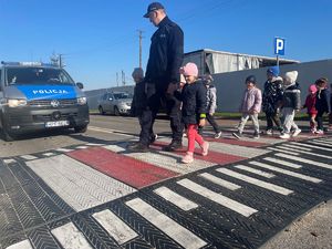 Dzieci trzymające odblaski w ręku, policjant przeprowadzający dzieci przez przejście dla pieszych.