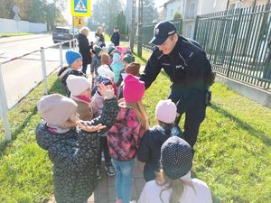 Dzieci trzymające odblaski w ręku, policjant przeprowadzający dzieci przez przejście dla pieszych.