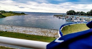 Widoczny w prawym dolnym rogu pagon na ramieniu, w tle port z łodziami.