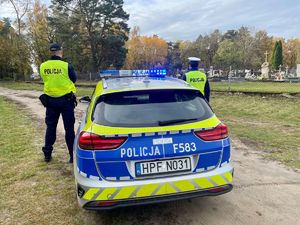 Policjanci przy radiowozie, w tle cmentarz.