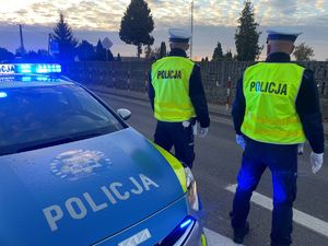 Policjanci przy radiowozie, w tle cmentarz.