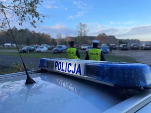 Policjanci przy radiowozie, w tle cmentarz.