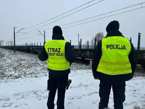 Policjant wraz z Funkcjonariuszem Straży Ochrony Kolei, w tle tory kolejowe.